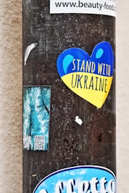 A vertical pole is pasted with various stickers. Prominently among them is a heart-shaped sticker in blue and yellow colors with the text 'Stand With Ukraine'. Additionally, there are other smaller stickers including a rectangular one with QR code and some partially ripped labels. The background is a neutral wall.