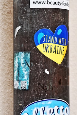 A vertical pole is pasted with various stickers. Prominently among them is a heart-shaped sticker in blue and yellow colors with the text 'Stand With Ukraine'. Additionally, there are other smaller stickers including a rectangular one with QR code and some partially ripped labels. The background is a neutral wall.