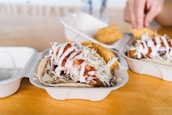 Close-up of a tray holding tacos filled with fried fish, shredded cabbage, and drizzled with white and red sauce. Part of a hand holding a tortilla chip can be seen in the background.