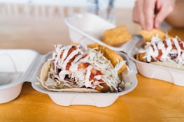 Close-up of a tray holding tacos filled with fried fish, shredded cabbage, and drizzled with white and red sauce. Part of a hand holding a tortilla chip can be seen in the background.