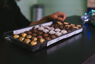A person is reaching towards a tray filled with a variety of chocolates. The chocolates are arranged neatly in rows, each with different colors and textures such as yellow, dark brown, and white with sprinkles. Labels in front of the tray indicate different flavors such as Passion Fruit and Dark & Stormy.