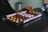 A person is reaching towards a tray filled with a variety of chocolates. The chocolates are arranged neatly in rows, each with different colors and textures such as yellow, dark brown, and white with sprinkles. Labels in front of the tray indicate different flavors such as Passion Fruit and Dark & Stormy.