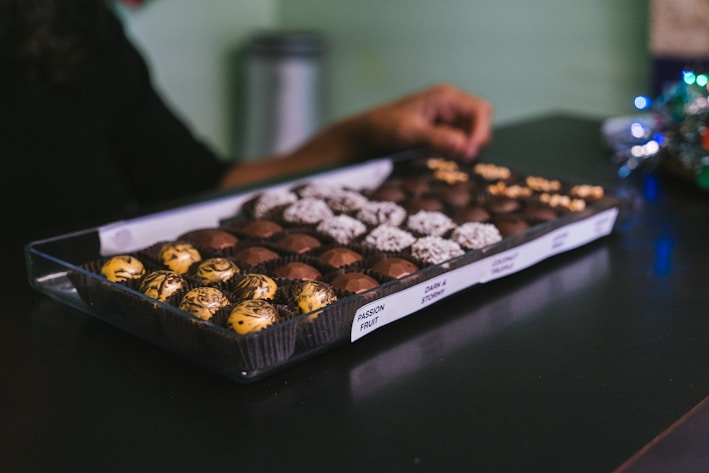A person is reaching towards a tray filled with a variety of chocolates. The chocolates are arranged neatly in rows, each with different colors and textures such as yellow, dark brown, and white with sprinkles. Labels in front of the tray indicate different flavors such as Passion Fruit and Dark & Stormy.