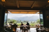 A cozy open-air dining area with wooden ceiling beams and tables set for meals, overlooking lush greenery and misty mountains in the distance. Potted plants add a touch of nature to the scene, while two people are seated at a table enjoying the view.