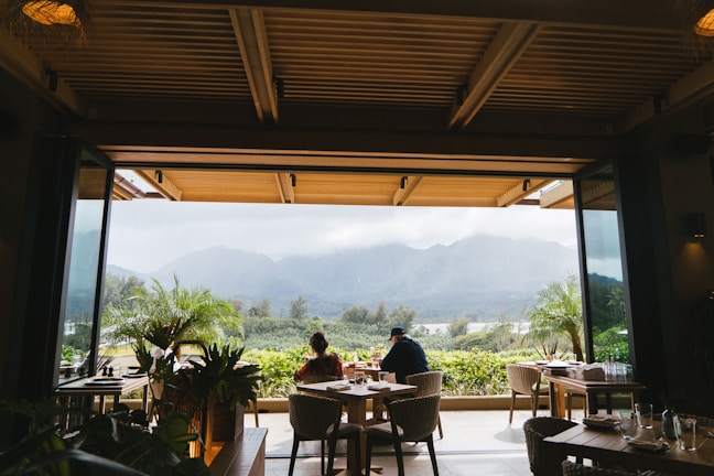 A cozy open-air dining area with wooden ceiling beams and tables set for meals, overlooking lush greenery and misty mountains in the distance. Potted plants add a touch of nature to the scene, while two people are seated at a table enjoying the view.