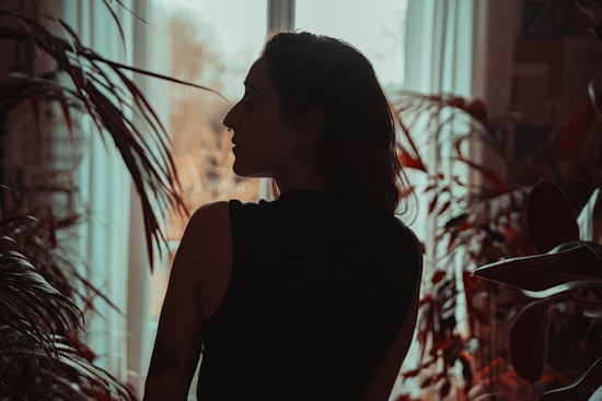 A person is silhouetted against a bright window, surrounded by lush plants and foliage. The mood is serene and introspective, with a focus on the interplay of light and shadow.