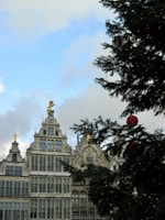 A large Christmas tree with red ornaments stands in the foreground, partially covering a portion of ornate, historic European buildings that feature intricate architectural details and statues on the rooftops. The sky is partly cloudy with patches of blue.