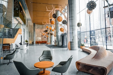 A modern and spacious lobby with large glass windows allowing natural light to flood the area. Hanging spherical lights in hues of orange and black add a contemporary feel. Sleek furnishings include a curved wooden bench and grey lounge chairs surrounding a small table. The space has a polished stone floor and an elegant reception desk near a staircase with glass railings.