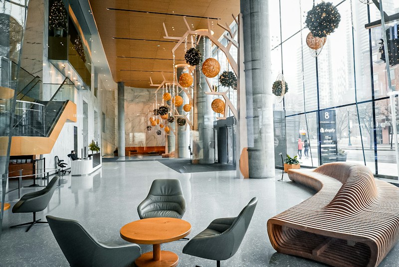 A modern and spacious lobby with large glass windows allowing natural light to flood the area. Hanging spherical lights in hues of orange and black add a contemporary feel. Sleek furnishings include a curved wooden bench and grey lounge chairs surrounding a small table. The space has a polished stone floor and an elegant reception desk near a staircase with glass railings.
