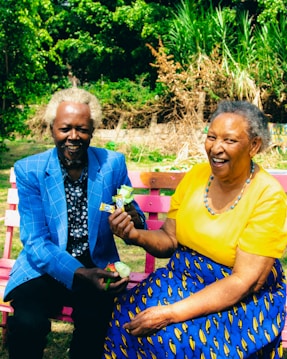 An elderly couple sits on a colorful pink garden bench surrounded by lush greenery. The woman is wearing a bright yellow top and a blue patterned skirt, smiling warmly while holding a flower. The man beside her is dressed in a blue checkered blazer with a dark patterned shirt underneath, also smiling while holding a small package. The background is filled with vibrant green plants and trees.