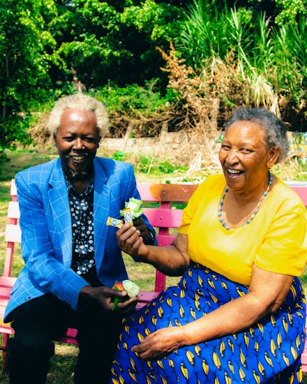 An elderly couple sits on a colorful pink garden bench surrounded by lush greenery. The woman is wearing a bright yellow top and a blue patterned skirt, smiling warmly while holding a flower. The man beside her is dressed in a blue checkered blazer with a dark patterned shirt underneath, also smiling while holding a small package. The background is filled with vibrant green plants and trees.