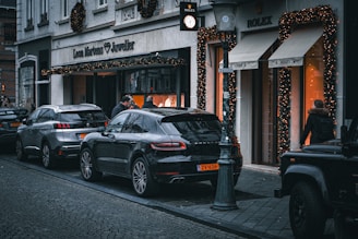A street scene featuring several parked cars along a cobblestone sidewalk. A jewelry store and a watch shop with festive decorations and illuminated displays are in the background. People are visible walking past or standing near the shop entrances.