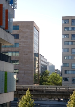 The image captures a modern urban scene with multiple high-rise buildings featuring large windows and varied architectural styles. The foreground includes a slightly elevated road or walkway, and a few green trees add a touch of nature to the scene. The sky appears clear, suggesting a sunny day.