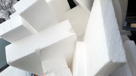 A cluster of white polystyrene foam blocks stacked in various angles. The blocks have a textured surface and appear to be used for insulation or packaging. The background includes parts of a car and some trees.