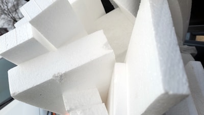 A cluster of white polystyrene foam blocks stacked in various angles. The blocks have a textured surface and appear to be used for insulation or packaging. The background includes parts of a car and some trees.
