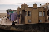 A seaside area features several quaint, colorful buildings with distinct architecture, including stucco walls and clay tile roofs. The scene includes a boardwalk and pier in the background, extending into a calm sea. People walk along the promenade, adding a leisurely and relaxed element to the setting.