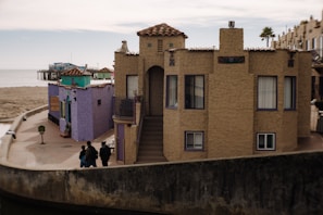 A seaside area features several quaint, colorful buildings with distinct architecture, including stucco walls and clay tile roofs. The scene includes a boardwalk and pier in the background, extending into a calm sea. People walk along the promenade, adding a leisurely and relaxed element to the setting.