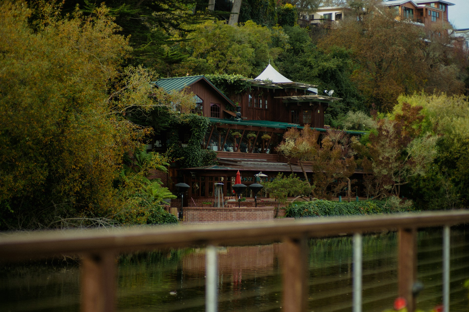 A rustic, multi-level wooden lodge is nestled within lush greenery, with ivy and trees surrounding it. The building's green roof blends with the natural environment. Patio umbrellas are visible on a terrace, suggesting an outdoor seating area. The foreground features a reflective body of water and a wooden railing.
