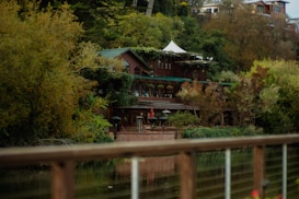 A rustic, multi-level wooden lodge is nestled within lush greenery, with ivy and trees surrounding it. The building's green roof blends with the natural environment. Patio umbrellas are visible on a terrace, suggesting an outdoor seating area. The foreground features a reflective body of water and a wooden railing.