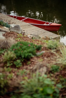 A red canoe is positioned against a wooden dock, with a serene body of water reflecting the surroundings. The foreground features lush greenery and rocks, partially out of focus.