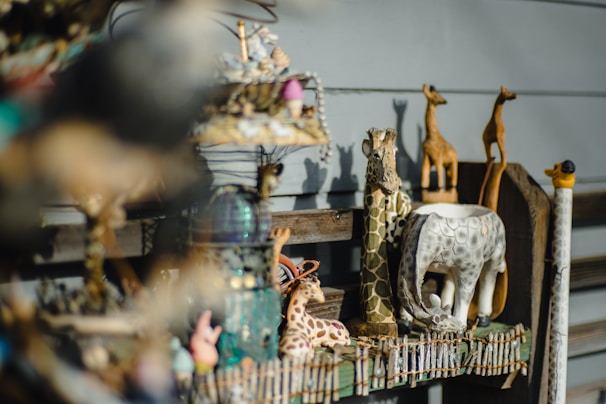 A collection of small, decorative animal figurines is displayed on a wooden shelf. The figurines include several giraffes and an elephant, crafted with intricate patterns and textures. The background is composed of wooden slats, and the focus on the objects creates a soft, blurred foreground effect.