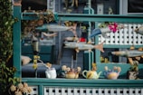 A decorative outdoor scene featuring a variety of colorful fish sculptures suspended in front of a wooden lattice fence. Below the fish, several teapot-shaped planters are arranged, each containing small succulents. The setup includes elements of nature and art, creating an eclectic and creative display.
