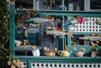 A decorative outdoor scene featuring a variety of colorful fish sculptures suspended in front of a wooden lattice fence. Below the fish, several teapot-shaped planters are arranged, each containing small succulents. The setup includes elements of nature and art, creating an eclectic and creative display.