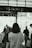 A monochrome photograph featuring a person with medium-length hair wearing a light-colored sweater and carrying a shoulder bag, standing amidst a bustling crowd inside a shopping or public area. The scene is filled with soft-focus and blurred silhouettes of people, giving a sense of motion and activity. There are bubbles floating throughout the space, adding a whimsical touch.