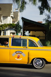 A classic yellow taxi with black and white checkered detailing is parked on the street. The taxi has a decorative miniature Christmas tree on its roof. The backdrop includes residential homes partially obscured by trees. The taxi has distinctive vintage styling, representing an older model vehicle.