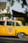 A classic yellow taxi with black and white checkered detailing is parked on the street. The taxi has a decorative miniature Christmas tree on its roof. The backdrop includes residential homes partially obscured by trees. The taxi has distinctive vintage styling, representing an older model vehicle.