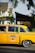 A classic yellow taxi with black and white checkered detailing is parked on the street. The taxi has a decorative miniature Christmas tree on its roof. The backdrop includes residential homes partially obscured by trees. The taxi has distinctive vintage styling, representing an older model vehicle.