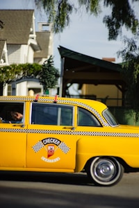 A classic yellow taxi with black and white checkered detailing is parked on the street. The taxi has a decorative miniature Christmas tree on its roof. The backdrop includes residential homes partially obscured by trees. The taxi has distinctive vintage styling, representing an older model vehicle.