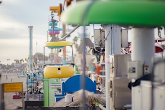 A colorful amusement park scene with a focus on a cable car system. The yellow and blue gondolas are hanging from a wire, moving past tall poles and various vibrant structures. In the background, the beach and other amusement park rides, like a vertical drop tower, are visible with palm trees dotting the landscape.