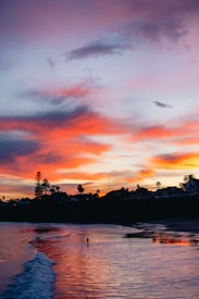 A beach scene at sunset, with vibrant colors painting the sky and reflecting on the water. Silhouettes of trees and houses can be seen on a distant shoreline. A lone figure is walking along the wet sand, adding a sense of scale and solitude.