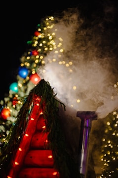 A festive scene featuring a wooden structure adorned with red lights and evergreen branches. In the background, a decorated Christmas tree glows with multicolored and warm white lights. A chimney emits swirling smoke, adding a mystical atmosphere.