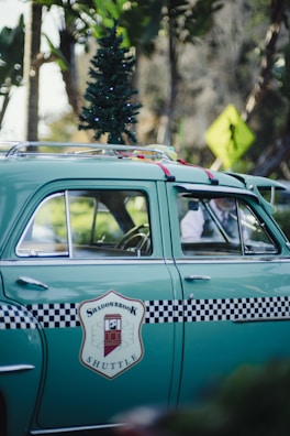 A vintage green car features a small Christmas tree on its roof and a checkered black and white design with a 'Shadowbrook Shuttle' logo on the side. The car is parked on a street, with tropical trees and a pedestrian crossing sign in the background.