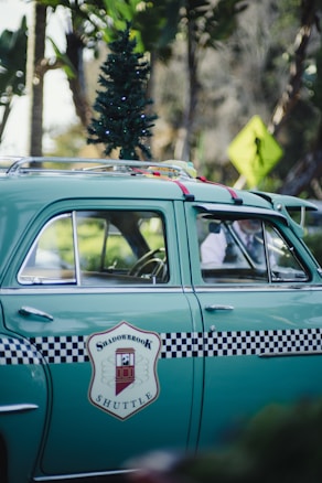A vintage green car features a small Christmas tree on its roof and a checkered black and white design with a 'Shadowbrook Shuttle' logo on the side. The car is parked on a street, with tropical trees and a pedestrian crossing sign in the background.