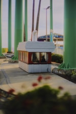 A small ticket booth or kiosk surrounded by tall green poles. The booth has a red bottom and white top, with large windows revealing a person sitting inside. Nearby are some flowerbeds with red flowers. In the background, there's a glimpse of a colorful awning, possibly part of a carnival or amusement park ride.