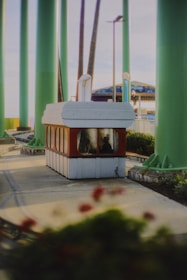 A small ticket booth or kiosk surrounded by tall green poles. The booth has a red bottom and white top, with large windows revealing a person sitting inside. Nearby are some flowerbeds with red flowers. In the background, there's a glimpse of a colorful awning, possibly part of a carnival or amusement park ride.