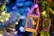 A small, decorative lantern hangs amid vibrant green foliage, containing a miniature fairy figurine with bright wings. The surroundings are illuminated with warm, colorful lights creating a whimsical and enchanting atmosphere.