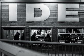 A monochromatic image capturing the side wall of a building with large letters 'IDE' prominently displayed. Through the windows below, silhouettes of people are seen, including a person wearing a hat. The interior is softly lit, revealing a decorated Christmas tree and festive wreaths, suggesting a cozy and inviting atmosphere.