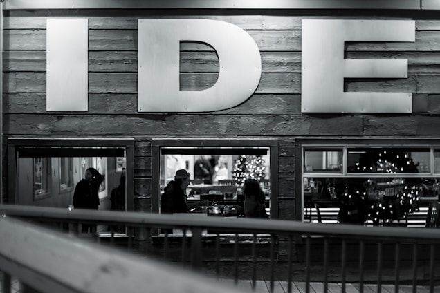 A monochromatic image capturing the side wall of a building with large letters 'IDE' prominently displayed. Through the windows below, silhouettes of people are seen, including a person wearing a hat. The interior is softly lit, revealing a decorated Christmas tree and festive wreaths, suggesting a cozy and inviting atmosphere.