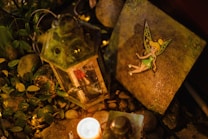 An enchanted garden scene featuring a glowing lantern, a small lit candle, surrounding greenery, and a decorative tile adorned with a fairy illustration. Pebbles and leaves are scattered throughout, creating a whimsical and magical atmosphere.