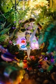 A whimsical fairy figurine sits amidst a vibrant, magical setting filled with colorful gemstones and lush greenery. Twinkling lights add a dreamlike quality to the scene, and the fairy wears a shimmering green dress and delicate wings.