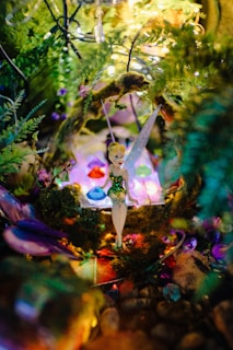 A whimsical fairy figurine sits amidst a vibrant, magical setting filled with colorful gemstones and lush greenery. Twinkling lights add a dreamlike quality to the scene, and the fairy wears a shimmering green dress and delicate wings.