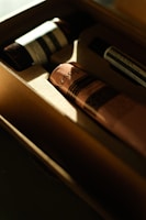 Several skincare products are placed inside a cardboard box, with sunlight creating a warm glow. The products have elegant, minimalist packaging, with one prominently visible pink tube labeled 'Aesop.' The lighting enhances the luxury aesthetic.