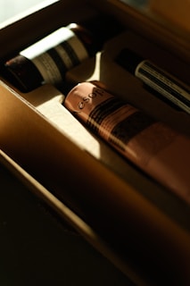 Several skincare products are placed inside a cardboard box, with sunlight creating a warm glow. The products have elegant, minimalist packaging, with one prominently visible pink tube labeled 'Aesop.' The lighting enhances the luxury aesthetic.