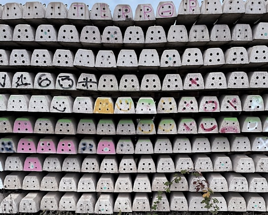 A wall of stacked, identical concrete blocks with circular and square cutouts, some featuring graffiti or colorful paint. The blocks vary in decoration, including smiley faces and abstract patterns. The bottom of the wall has some foliage growing against it.