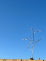 A clear blue sky serves as the backdrop to a rooftop-mounted antenna system, featuring multiple horizontal and vertical elements. The structure is built of metal and stands prominently against the sky. A beige-colored wall is visible at the bottom of the image.