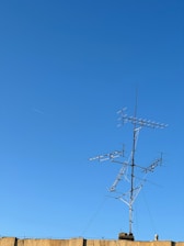 A clear blue sky serves as the backdrop to a rooftop-mounted antenna system, featuring multiple horizontal and vertical elements. The structure is built of metal and stands prominently against the sky. A beige-colored wall is visible at the bottom of the image.